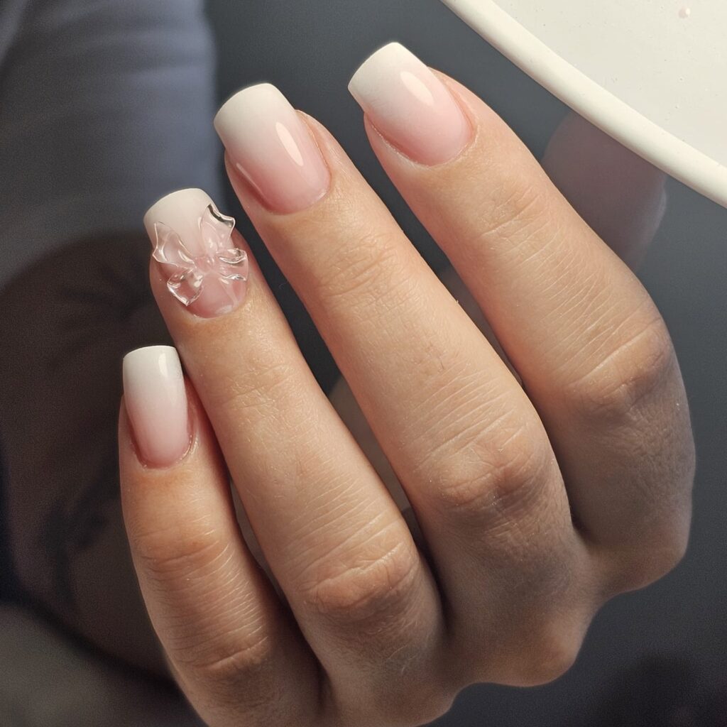 Ongles forme "square" avec un dégradé de couleur claires, rose à partir des cuticules qui tourne au blanc à l'extrémité des ongles. Sur l'annulaire, un noeud transparent est déposé au milieu de l'ongle.