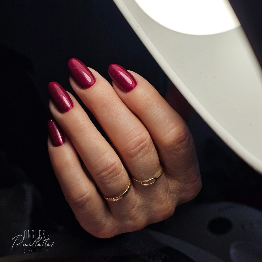 Ongles en forme d'amande, avec une pose de vernis semi-permanent bordeaux pailleté.