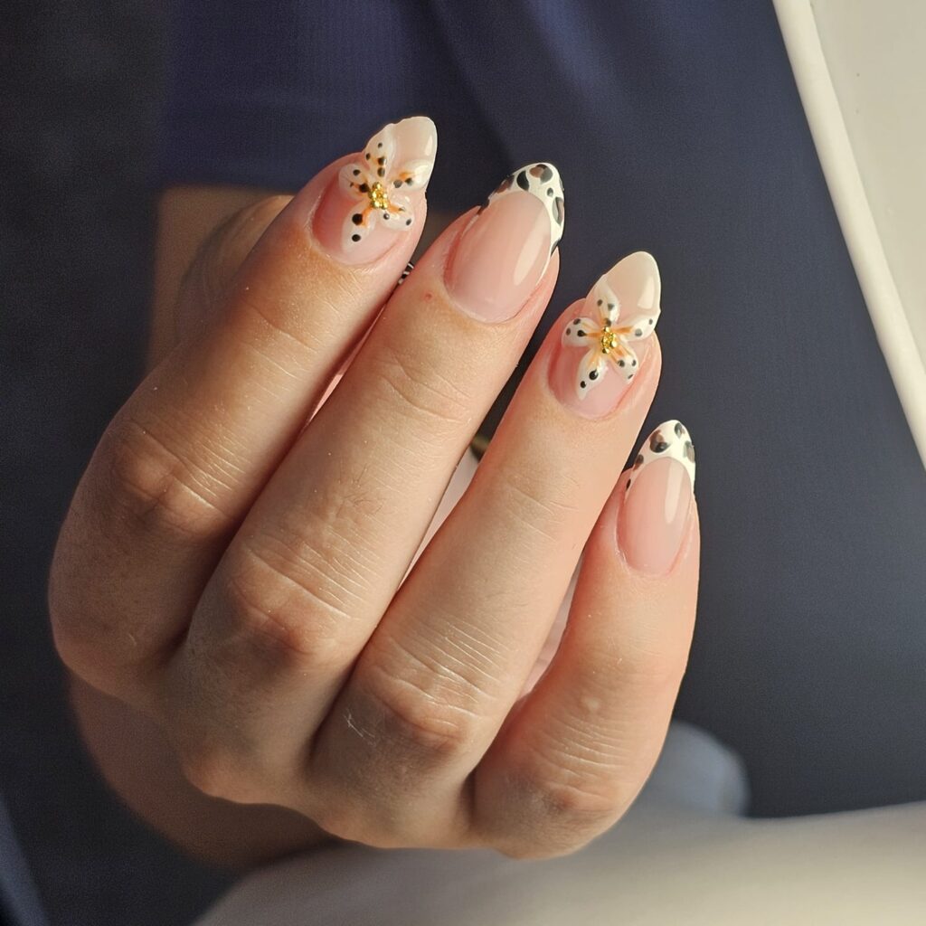 Pose de nail art. Des ongles rosés avec des motifs de vache aux extrémités du pouce, du majeur et de l'auriculaire. Ensuite sur l'index et l'annulaire, il y a une fleur, qui elles aussi sont parsemées de petites tâches noires.