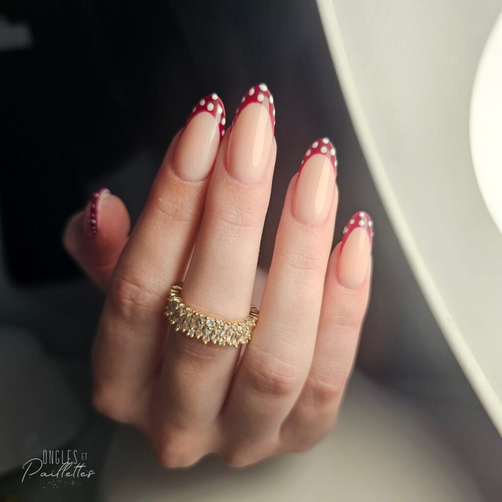Ongles en frome d'amande, avec une pose de vernis semi-permanent rose nude. L'extrémité des doigts est rouge bordeaux avec des points blancs.