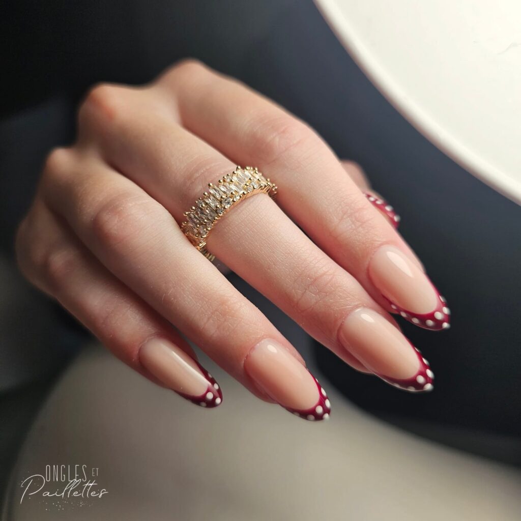 Ongles en frome d'amande, avec une pose de vernis semi-permanent rose nude. L'extrémité des doigts est rouge bordeaux avec des points blancs.